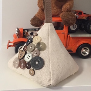 May include: A beige triangular doorstop with a brown fabric handle. The doorstop is decorated with a variety of buttons in different colours and patterns, including some with the text "SUKO CARGO" printed on them.