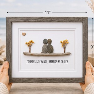 May include: A framed pebble art piece measuring 11 inches by 9 inches. It features two figures made of smooth gray stones sitting on a piece of driftwood, flanked by yellow dried flowers. A small wooden heart and the text COUSINS BY CHANCE, FRIENDS BY CHOICE are included.