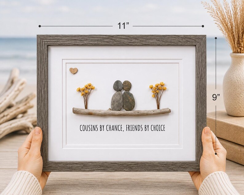May include: A framed pebble art piece measuring 11 inches by 9 inches. It features two figures made of smooth gray stones sitting on a piece of driftwood, flanked by yellow dried flowers. A small wooden heart and the text COUSINS BY CHANCE, FRIENDS BY CHOICE are included.
