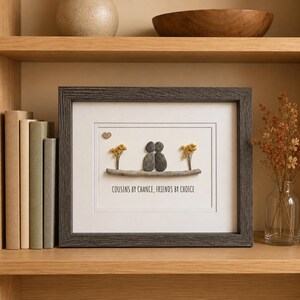 May include: A framed wall art piece featuring pebble art of two figures sitting on a branch with yellow dried flowers and a small heart. The text reads COUSINS BY CHANCE, FRIENDS BY CHOICE. The frame is dark gray with a textured wood grain finish.