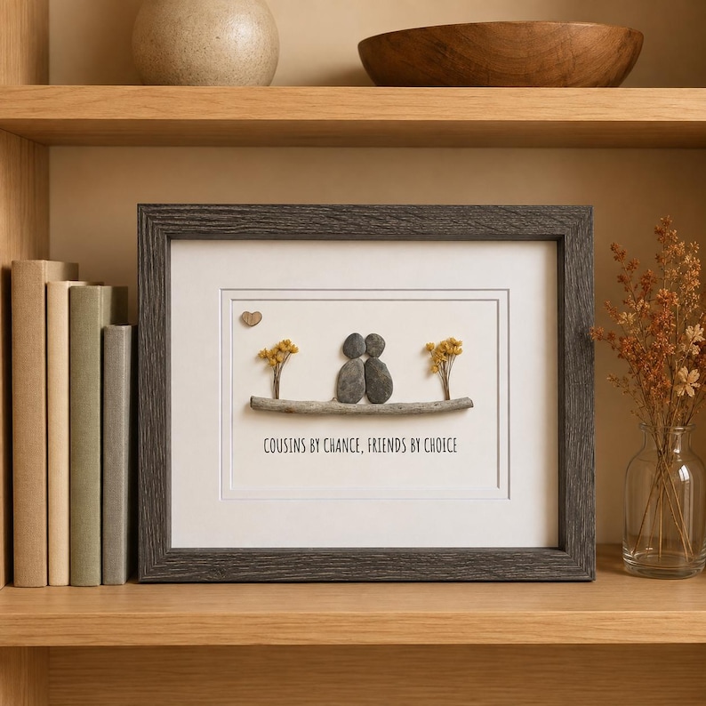 May include: A framed wall art piece featuring pebble art of two figures sitting on a branch with yellow dried flowers and a small heart. The text reads COUSINS BY CHANCE, FRIENDS BY CHOICE. The frame is dark gray with a textured wood grain finish.