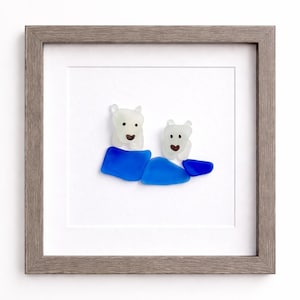 Puede incluir: Obra de arte enmarcada con dos figuras de oso polar hechas de vidrio marino blanco y azul. Los osos est&aacute;n sentados sobre vidrio azul, con un marco de madera gris. La obra est&aacute; sobre un fondo blanco.