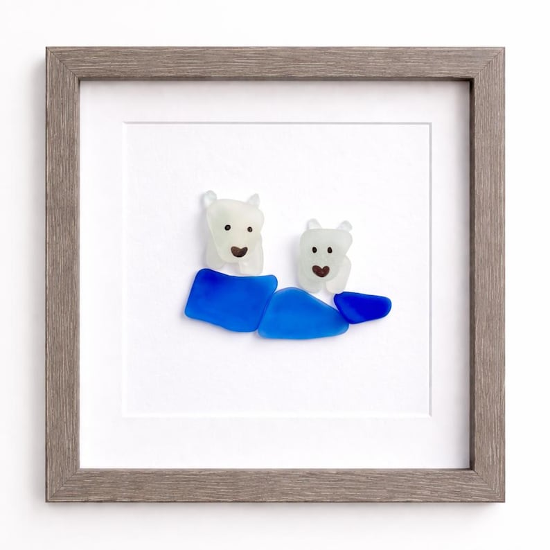 Puede incluir: Obra de arte enmarcada con dos figuras de oso polar hechas de vidrio marino blanco y azul. Los osos est&aacute;n sentados sobre vidrio azul, con un marco de madera gris. La obra est&aacute; sobre un fondo blanco.