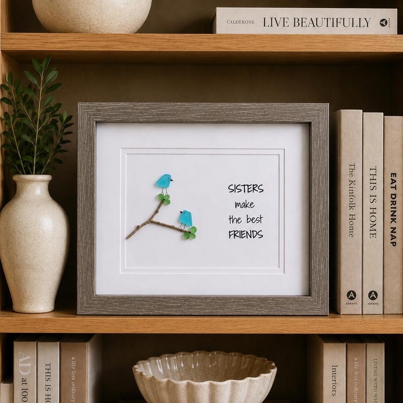 May include: A framed wall art piece featuring two light blue sea glass birds perched on a small brown twig with green sea glass leaves. The white background displays the black text: SISTERS make the best FRIENDS. The frame is textured gray wood.