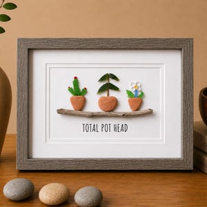 Peut inclure: Une œuvre d'art murale encadrée représentant trois petites plantes en pot faites de galets et de brindilles reposant sur une branche horizontale. Les plantes incluent un cactus, une plante à feuilles et une fleur. Le texte TOTAL POT HEAD est inscrit.