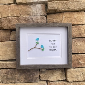 May include: A framed wall art piece featuring two blue birds crafted from sea glass perched on a small brown twig with green leaves. The white background displays the black text: SISTERS make the best FRIENDS. The frame is dark gray wood.