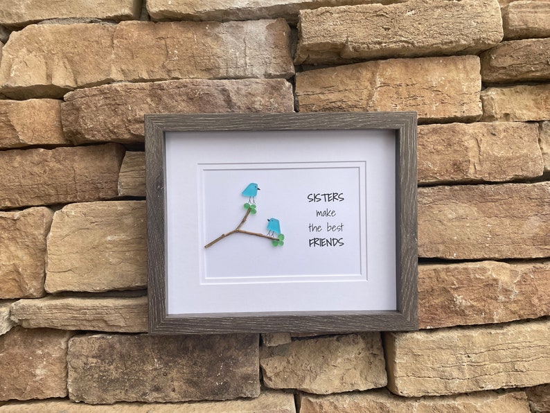 May include: A framed wall art piece featuring two blue birds crafted from sea glass perched on a small brown twig with green leaves. The white background displays the black text: SISTERS make the best FRIENDS. The frame is dark gray wood.