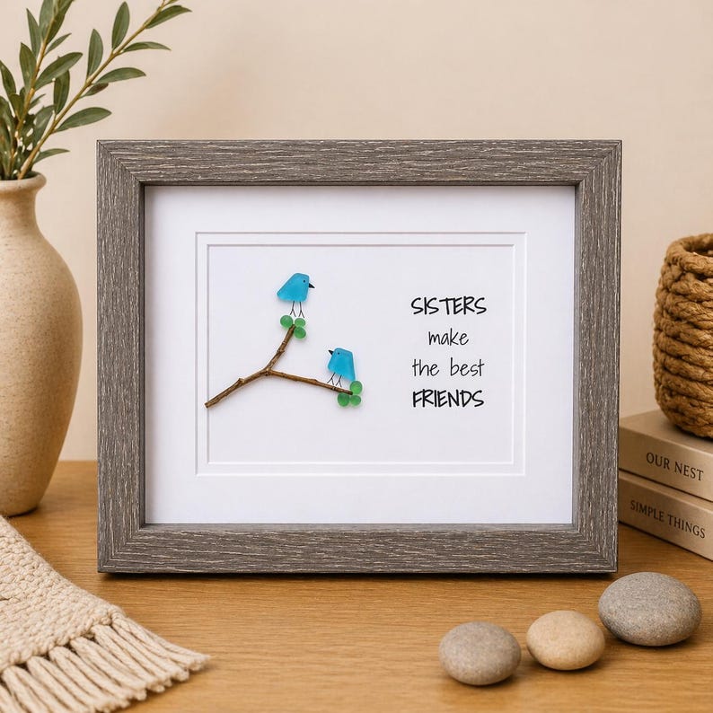 May include: A framed wall art piece featuring two blue sea glass birds perched on a brown twig with green sea glass leaves. The white background displays the black text: SISTERS make the best FRIENDS. The frame is a textured, dark gray wood finish.
