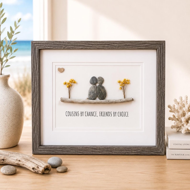 May include: A framed wall art piece featuring two pebble figures sitting on a piece of driftwood, flanked by small yellow dried flowers. A small wooden heart is positioned above the figures. The text reads: COUSINS BY CHANCE, FRIENDS BY CHOICE.