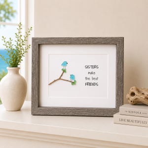 May include: A framed wall art piece featuring two light blue sea glass birds perched on a brown twig with small green sea glass leaves. The white background displays the black text: SISTERS make the best FRIENDS. The frame has a grey wood grain finish.