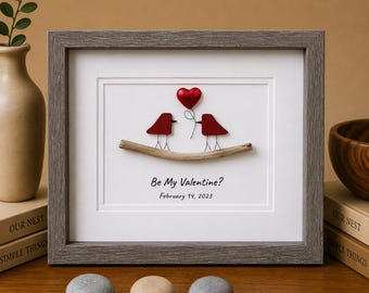 May include: A framed wall art piece featuring two red bird silhouettes perched on a piece of driftwood. A red heart is attached to one bird by a thin wire. The text "Be My Valentine?" and "February 14, 2023" is printed below the birds.