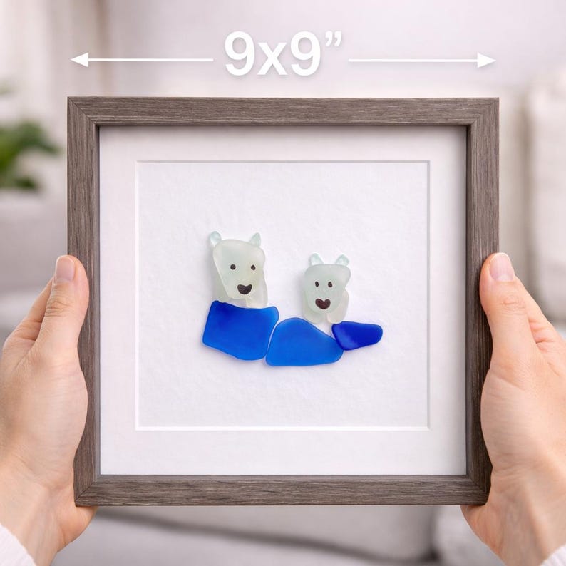 Puede incluir: Una obra de arte enmarcada con dos figuras de osos polares hechas de vidrio marino, con detalles azules. El marco es gris y mide 23x23 cm.