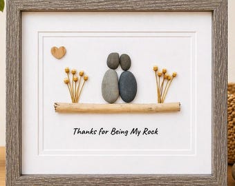 May include: A framed pebble art piece featuring two figures made of grey and dark grey stones standing on a horizontal piece of driftwood. The artwork includes dried yellow flowers, a small wooden heart, and the text Thanks for Being My Rock.