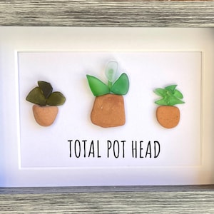 May include: A framed piece of art featuring three potted plants made from sea glass. The plants are green and brown and the pots are brown. The text "TOTAL POT HEAD" is written below the plants.