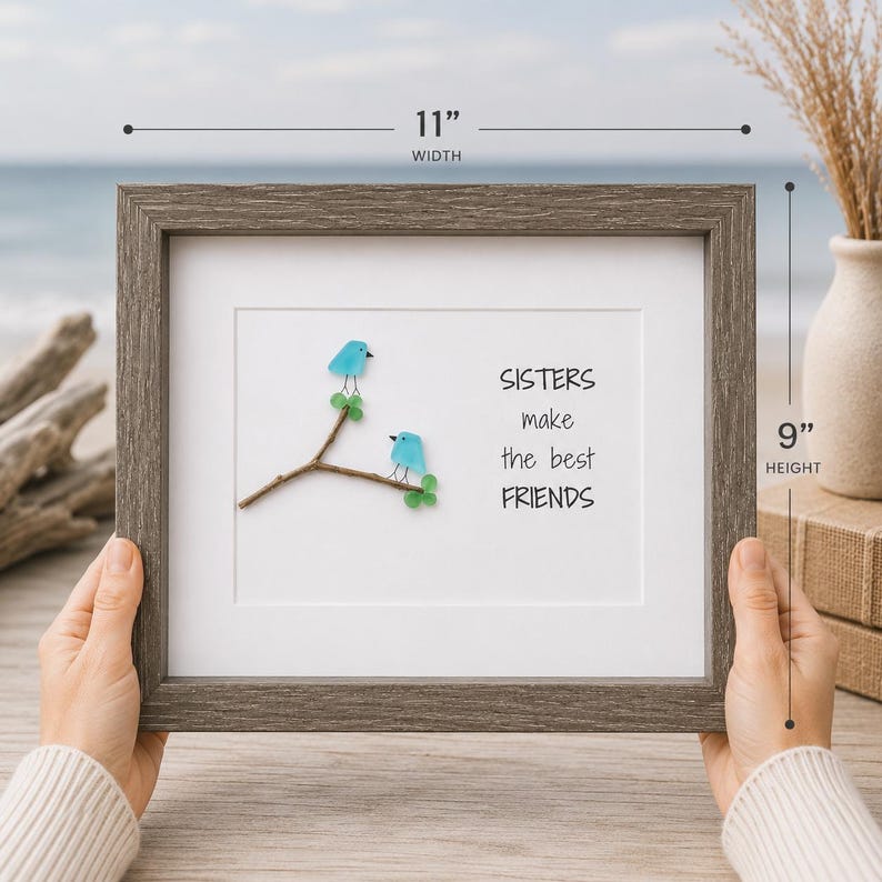 May include: A framed wall art piece featuring two blue sea glass birds perched on a brown twig with green glass leaves. The text reads SISTERS make the best FRIENDS. The frame is dark gray wood, measuring 11 inches in width and 9 inches in height.