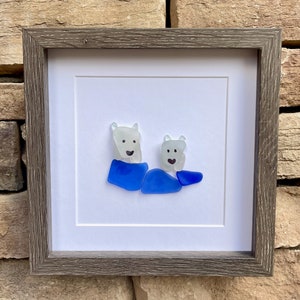 Puede incluir: Obra de arte enmarcada con un dise&ntilde;o de dos osos polares de vidrio de mar. Los osos est&aacute;n hechos de vidrio blanco transl&uacute;cido con ojos y sonrisas de puntos negros. Llevan ropa de vidrio azul. La obra est&aacute; enmarcada en un marco de madera gris.