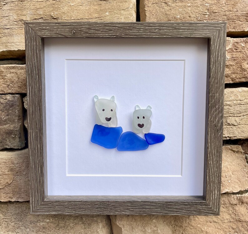 Puede incluir: Obra de arte enmarcada con un dise&ntilde;o de dos osos polares de vidrio de mar. Los osos est&aacute;n hechos de vidrio blanco transl&uacute;cido con ojos y sonrisas de puntos negros. Llevan ropa de vidrio azul. La obra est&aacute; enmarcada en un marco de madera gris.