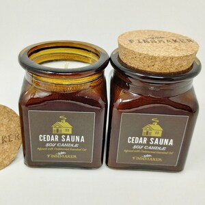 May include: Two amber glass jars with cork lids containing soy candles. The labels are brown with gold lettering and a small cabin illustration. The text on the labels reads "Cedar Sauna Soy Candle Infused with Cedarwood Essential Oil Finnmaker".