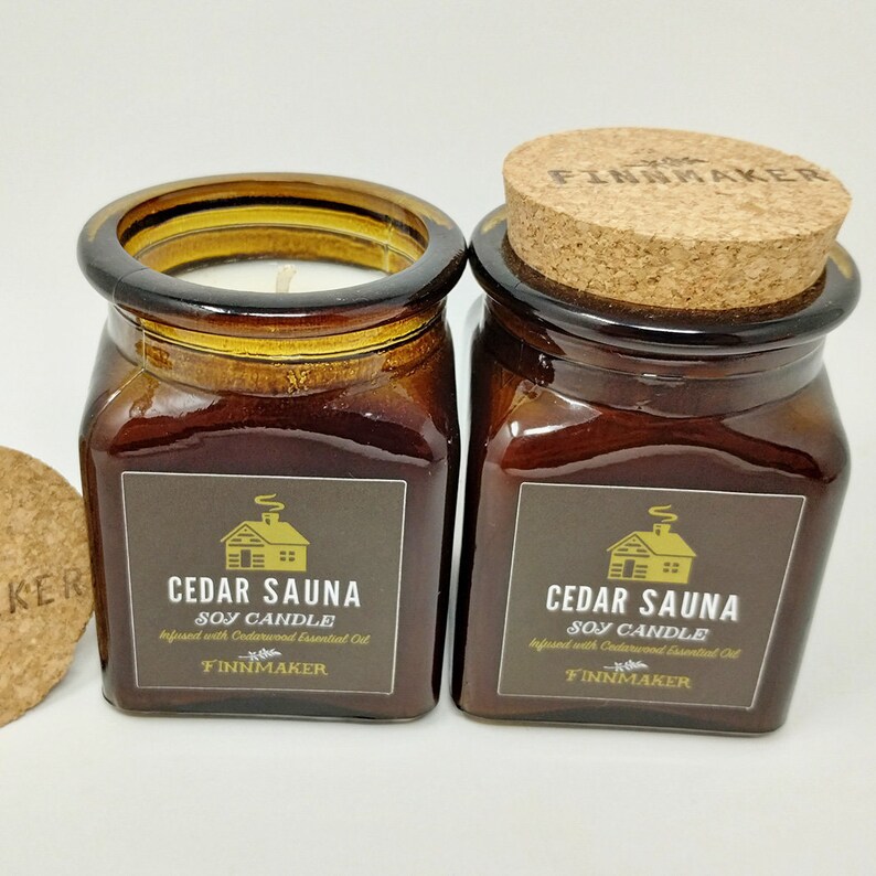 May include: Two amber glass jars with cork lids containing soy candles. The labels are brown with gold lettering and a small cabin illustration. The text on the labels reads "Cedar Sauna Soy Candle Infused with Cedarwood Essential Oil Finnmaker".