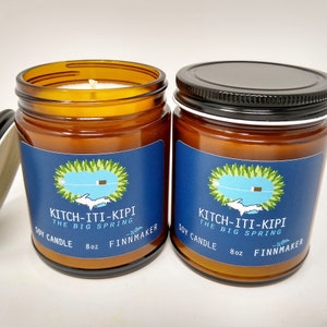 May include: Two amber glass jars with black lids and blue labels. The labels have a white outline of Michigan with a small cabin on a lake. The text on the labels reads "Kitch-iti-kipi The Big Spring Soy Candle 8oz Finnmaker".