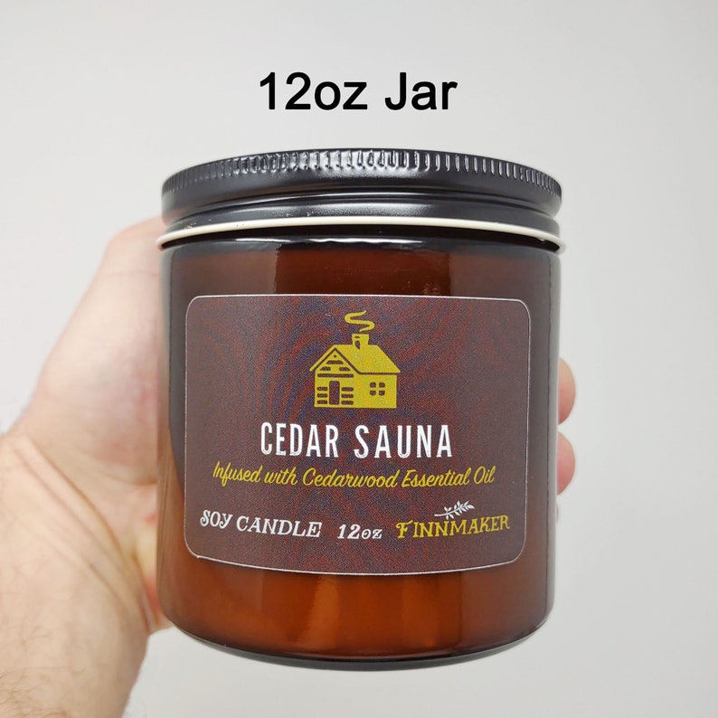 May include: A 12oz amber glass jar candle with a black lid. The label is brown with a gold cabin illustration and the text "CEDAR SAUNA Infused with Cedarwood Essential Oil SOY CANDLE 12oz FINNMAKER".