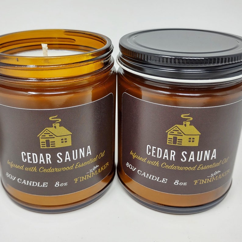 May include: Two amber glass jars with black lids containing soy candles. The labels are brown with gold lettering and a cabin illustration. The text on the labels reads "Cedar Sauna Infused with Cedarwood Essential Oil Soy Candle 8oz Finnmaker".