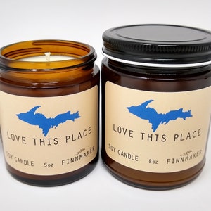 May include: Two amber-colored glass jars of soy candles. Each jar has a label with a blue outline of a landmass and the text "LOVE THIS PLACE". One jar is 5 oz, the other is 8 oz. Both jars have the text "SOY CANDLE" and "FINNMAKER".