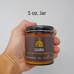 May include: A 5 oz. amber glass jar candle with a black lid. The label is brown with gold text and a graphic of a cabin with smoke coming from the chimney. The text on the label reads "SAUNA infused with Cedarwood Essential Oil SOY CANDLE 5oz FINNMAKER".