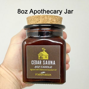 May include: An 8 ounce apothecary jar candle with a cork lid. The label is brown with a yellow cabin illustration and the text "CEDAR SAUNA SOY CANDLE Infused with Cedarwood Essential Oil FINNMAKER".