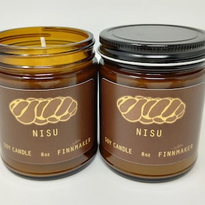 Pode incluir: Dois frascos de vidro castanho com tampas pretas contendo velas de soja. Os rótulos são castanhos com um desenho de pão trançado amarelo e o texto "Nisu Soy Candle 8oz Finnmaker".