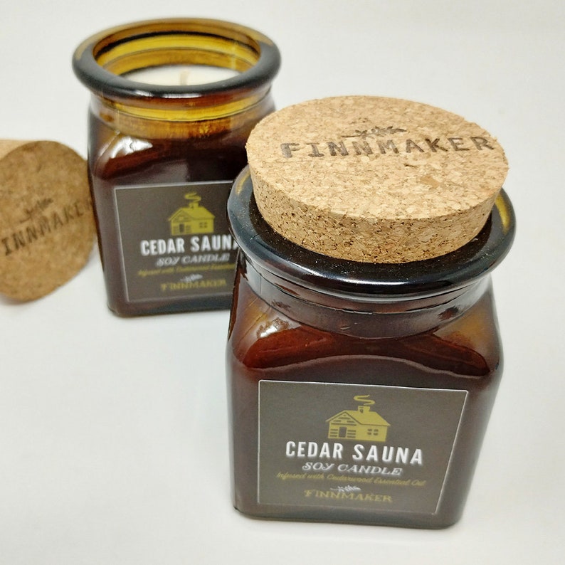 May include: Two amber glass jars with cork lids. The jars contain soy candles with a cedar sauna scent. The labels are black with white text and a small cabin icon. The text on the label reads "Cedar Sauna Soy Candle Infused with Cedarwood Essential Oil Finnmaker".