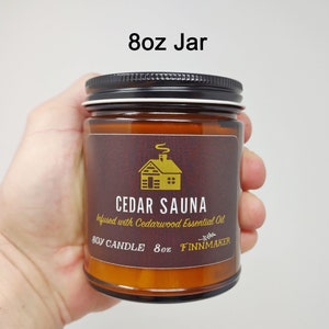May include: An 8 oz amber glass jar candle with a black lid. The label is brown with gold lettering and a gold cabin illustration. The text on the label reads "Cedar Sauna Infused with Cedarwood Essential Oil Soy Candle 8oz Finnmaker".
