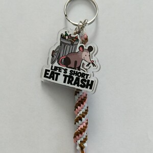May include: A keychain with a brown, white, and pink braided cord. The keychain charm is a clear plastic circle with a cartoon opossum eating trash from a garbage can. The text on the charm reads "Life's Short Eat Trash".