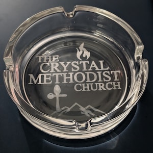 May include: A clear glass ashtray with an etched design. The design includes a flame, the words "The Crystal Methodist Church", a cross, and mountains.