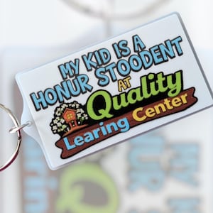 May include: A rectangular keychain with the text "MY KID IS A HONOR STOODENT AT Quality Learing Center" in blue, green, and brown. The keychain has a silver ring attached.