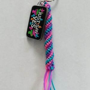 May include: A keychain with a silver ring and a woven cord in shades of pink, purple, and teal. The keychain has a rectangular charm with the words "JUST SAY YES" and colorful leaf designs on a black background.