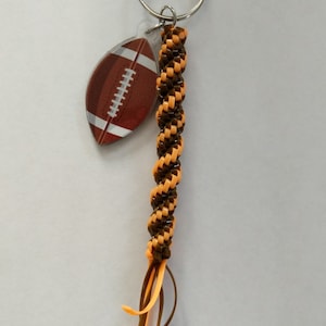May include: A brown and orange braided keychain with a football-shaped charm. The charm is clear plastic with a brown football design.