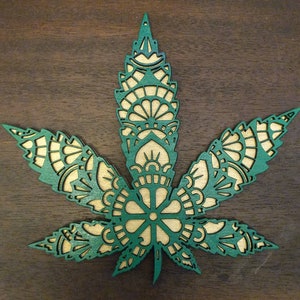May include: A green and brown wooden mandala design in the shape of a marijuana leaf. The design is intricate and detailed, with a variety of shapes and patterns.