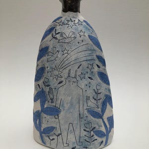 May include: A ceramic bottle with a dark rim and a light blue body. The bottle is decorated with blue leaves, stars, birds, and a figure with horns. The design is hand-drawn and whimsical.
