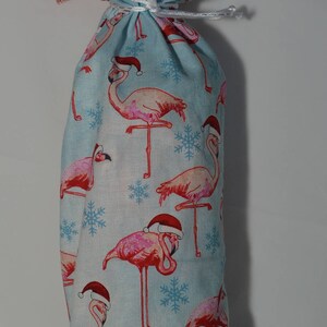 May include: A blue fabric wine bottle bag with a red and white snowflake pattern and pink flamingos wearing Santa hats.