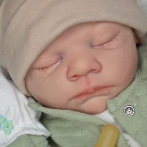 May include: A realistic reborn doll with closed eyes, wearing a beige knit cap and a sage green quilted jacket with a silver snap button. The doll has a pale complexion, visible skin texture, and is dressed in a white garment with small colorful patterns.