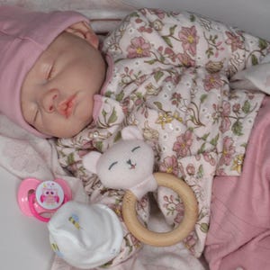 May include: A sleeping baby doll wearing a pink beanie and a floral long-sleeved top with matching pink pants. The doll is wrapped in a floral blanket and holds a wooden ring rattle with a plush animal head, accompanied by a pink pacifier.