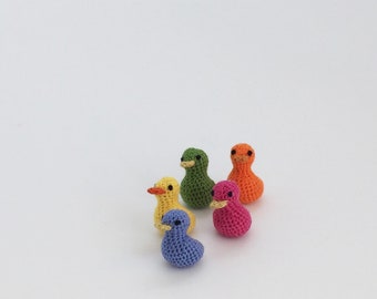 May include: A collection of six small, crocheted ducklings in various colors: yellow, green, orange, pink, blue, and yellow. Each duckling has a small beak and a simple, rounded shape. The ducks are arranged on a white surface.