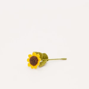 May include: A small crocheted sunflower with a brown center and yellow petals, attached to a green stem with leaves.