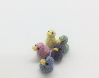 May include: Six small, crocheted ducklings in various pastel colors: yellow, pink, green, purple, and blue. Each duckling has a yellow beak and black eyes. The ducks are arranged on a white surface.