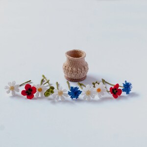 May include: A crocheted flower arrangement with white daisies, red poppies, and blue cornflowers. The flowers are arranged around a small crocheted basket.