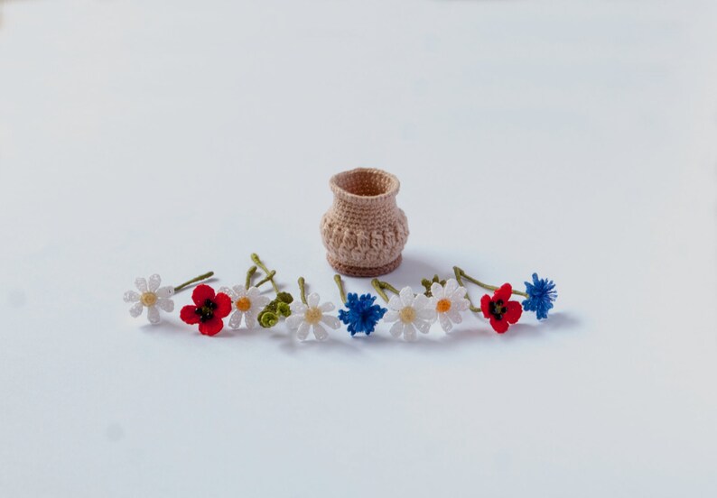 May include: A crocheted flower arrangement with white daisies, red poppies, and blue cornflowers. The flowers are arranged around a small crocheted basket.