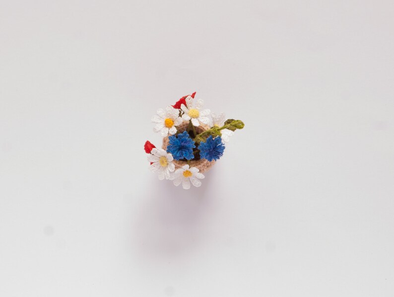 May include: A small crocheted bouquet of white daisies and blue cornflowers in a brown burlap pot.