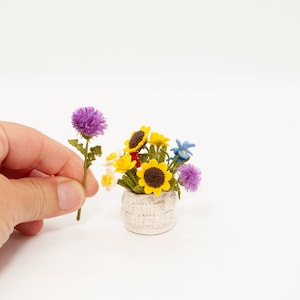 May include: A crocheted flower arrangement in a white crocheted pot. The arrangement includes yellow sunflowers, purple thistles, and white flowers.