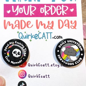 May include: Two black button pins with white text and graphics. The first pin says "Dead Ass Fucking Tired" with a skull and rainbow. The second pin says "Daily Shitstorm Survivors Club" with a gray cloud and rainbow. The pins are on a white card with the text "Thank You Your Order Made My Day QuirkeCATT.com" and social media handles.